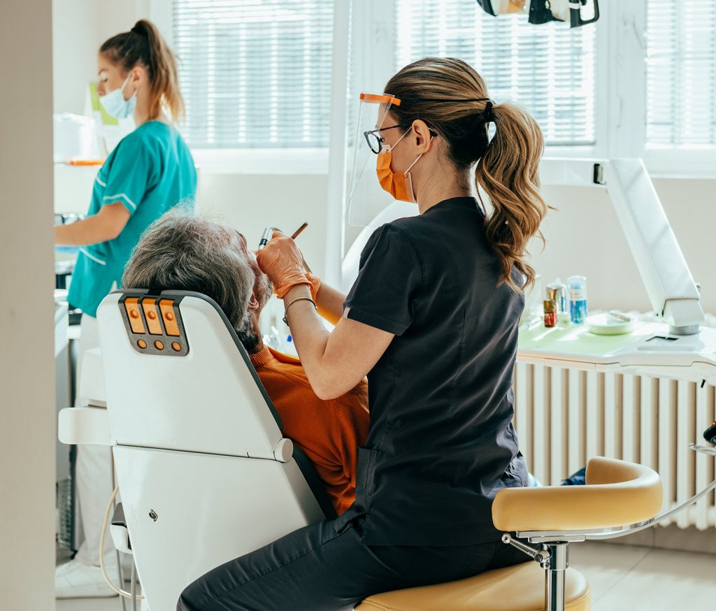 Frequently Asked Questions 1 Dentist in a face shield treating a patient while an assistant preps tools at our NYC dental clinic